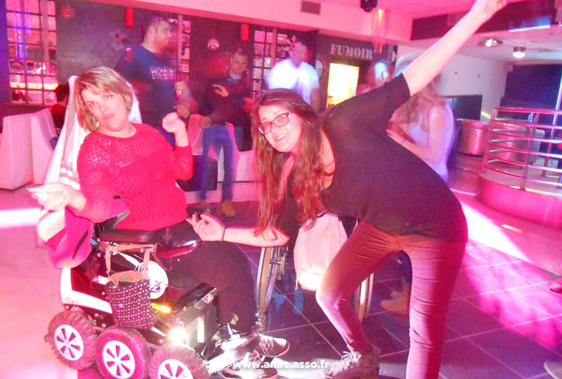 Une jeune femme en fauteuil roulant électrique et une accompagnatrice en train de danser lors d'une soirée discothèque
