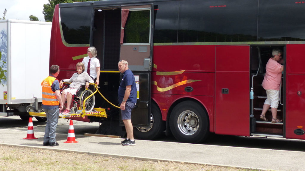 Une dame en fauteuil roulant descend d'un car adapté grâce à une plateforme élévatrice. Voyage et handicap Anaé