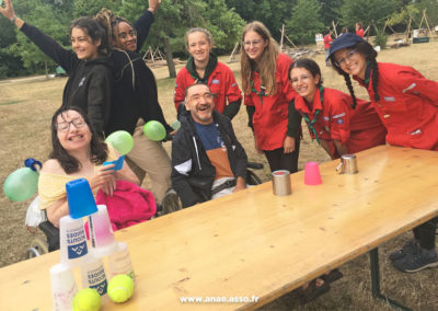 Séjour adapté PMR handicap Anaé Vacances à Jambville l'été 2022. Des vacanciers en fauteuil roulant et leurs animateurs prennent une photo de groupe avec de jeunes scouts qui ont organisé pour eux un jeu de piste.