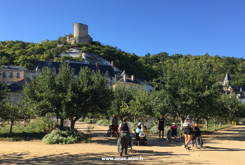 Séjour adapté PMR handicap Anaé Vacances à Jambville l'été 2022. Des vacanciers en fauteuil roulant et leurs animateurs se promènent dans un parc. Vue sur le château de la Roche-Guyon.