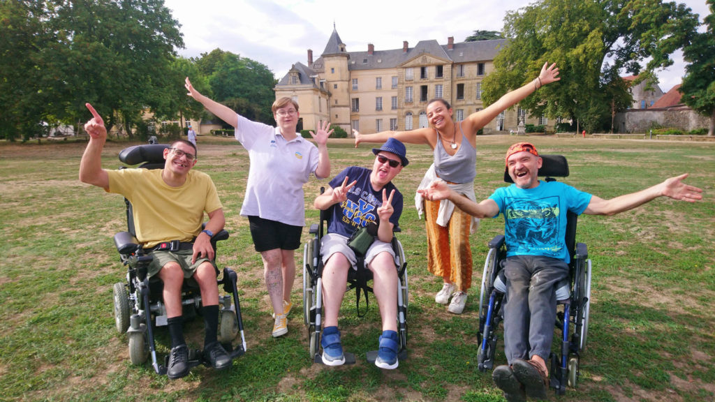 Séjour adapté handicap moteur avec Anaé Vacances au Château de Jambville en région parisienne. Des vacanciers heureux lèvent les mains en l'air devant le château.