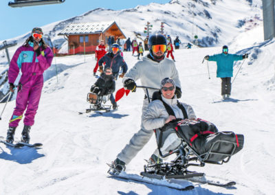 Séjour adapté handicap hiver 2023 avec Anaé Vacances. Une femme à mobilité réduite est heureuse de faire du ski assis avec un moniteur.