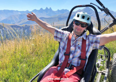 Activité adaptée PMR à la montagne. Un vacancier à mobilité réduite prend la pose dans un fauteuil tout terrain appelé Cimgo. Il est heureux et il lève les bras vers le ciel.
