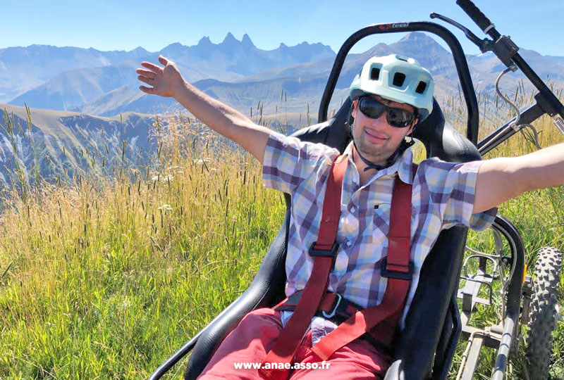 Activité adaptée PMR à la montagne. Un vacancier à mobilité réduite prend la pose dans un fauteuil tout terrain appelé Cimgo. Il est heureux et il lève les bras vers le ciel.