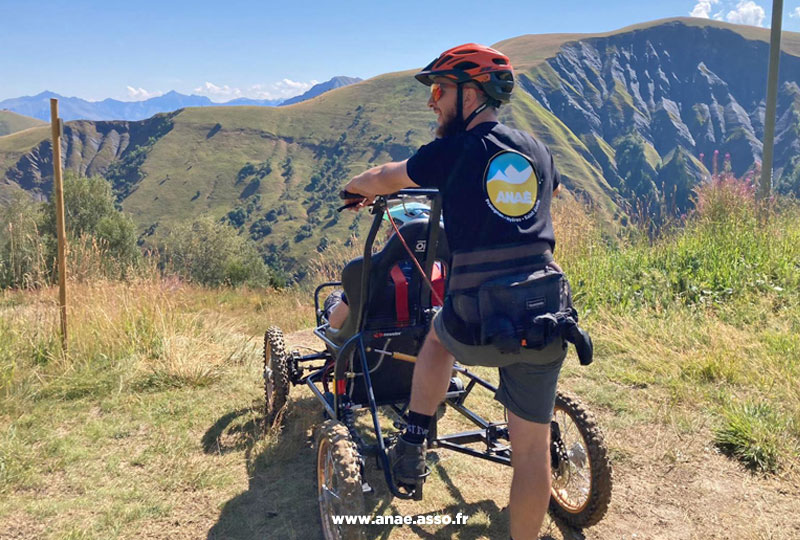 Activité adaptée PMR à la montagne. Un vacancier à mobilité réduite et son accompagnateur prennent la pose dans un fauteuil tout terrain appelé Cimgo