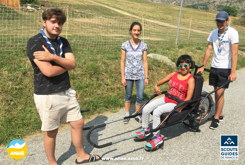 Activité adaptée PMR à la montagne. Des jeunes scouts portent une femme handicapée moteur lors d'une excursion en joëlette en Savoie.