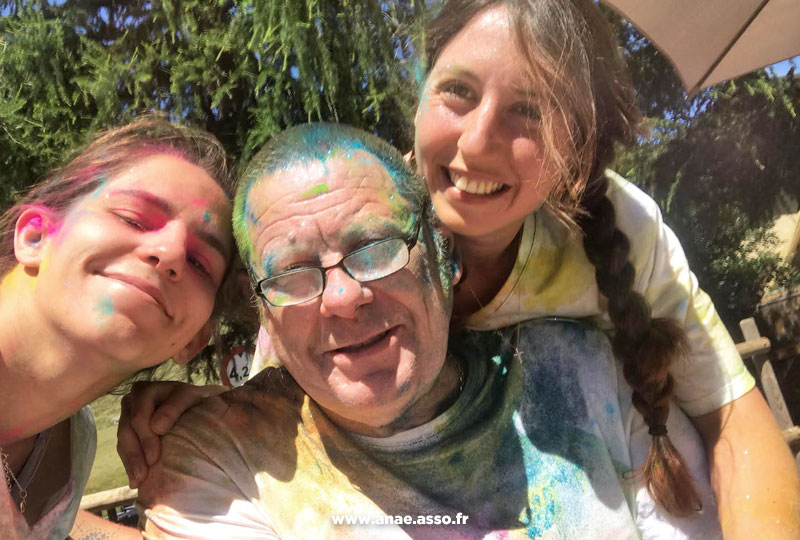 sejour-adapte-handicap-moteur-pmr-anae-vacances-pralognan-vanoise-savoie-vacancier-aidant-color-run-2022 Un vacancier en fauteuil roulant et deux animatrices de séjour adapté lors d'une color run. Ils ont le visage coloré.