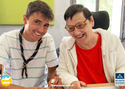 Une femme en fauteuil roulant fait une activité dessin avec un scout lors d'un séjour adapté PMR avec Anaé Vacances. Ils sourient tous les deux.