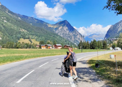 Une animatrice de séjour adapté pousse le fauteuil roulant d'un vacancier handicapé moteur. Vue sur les montagnes de Pralognan la Vanoise en Savoie.