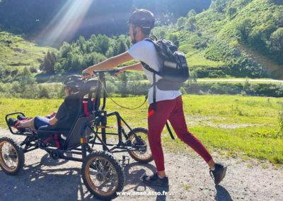Séjour de vacances adaptées pour personnes en situation de handicap moteur. Un vacancier se promène avec son accompagnateur à la montagne en cimgo, un fauteuil de descente tout terrain.