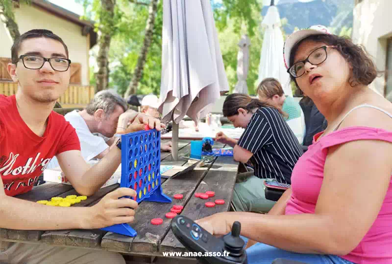 Séjour adapté handicap à la montagne. Une jeune femme en fauteuil roulant fait joue avec un animateur sur une table en terrasse.